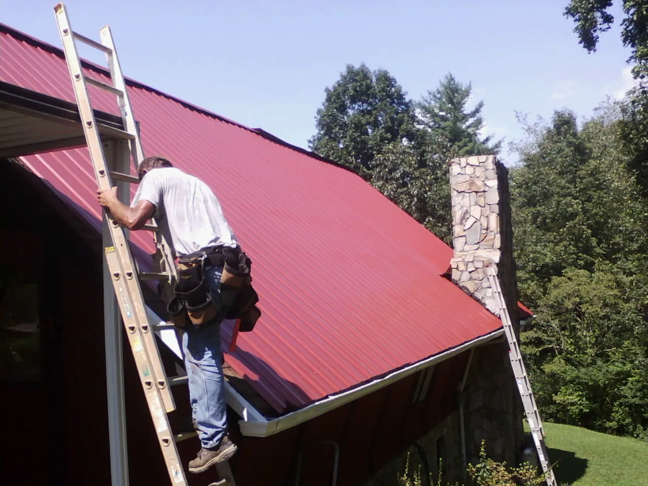 Roofer on a ladder beside a red metal roof and stone chimney — Soffit & Fascia Repair by Roofing Brothers in Krum